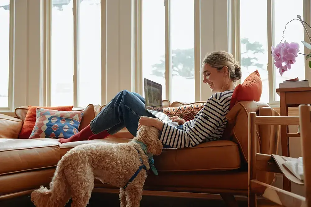Femme assise sur un canapé avec un chien à ses pieds, travaillant sur un ordinateur portable près d'une fenêtre lumineuse.